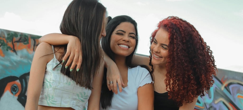 Three young women smiling and embracing outdoors, reflecting connection and support within an inclusive, identity-affirming environment