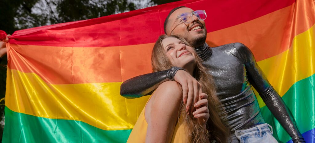Two friends smiling and embracing in front of a rainbow flag, representing connection, community, identity, and comfort within conversations around intimacy and anxiety challenges