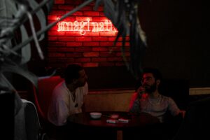 Two people sitting together in a dimly lit lounge beneath a red neon sign