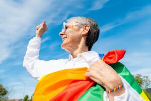 Older LGBTQIA+ adult smiling while holding a Pride flag, representing the experience and joy of coming out later in life.